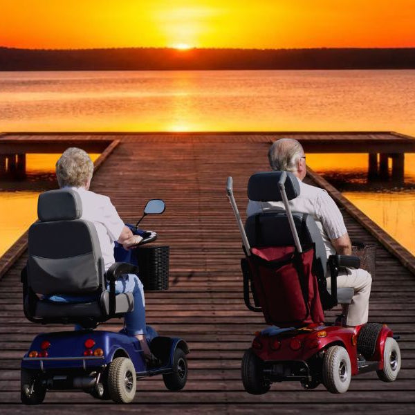 Two people riding mobility scooters side by side down a lakeside boardwalk toward a sunset