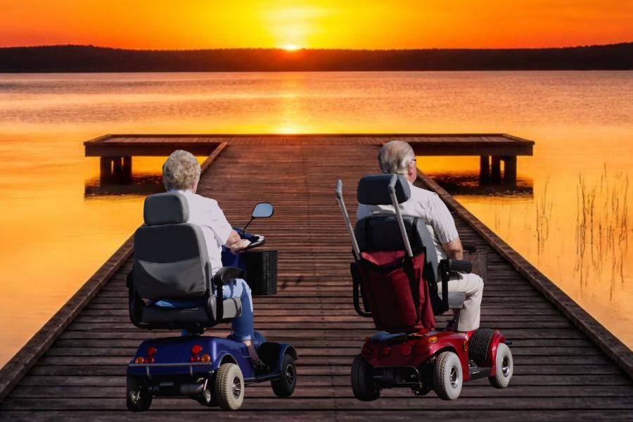 Two people riding mobility scooters side by side down a lakeside boardwalk toward a sunset