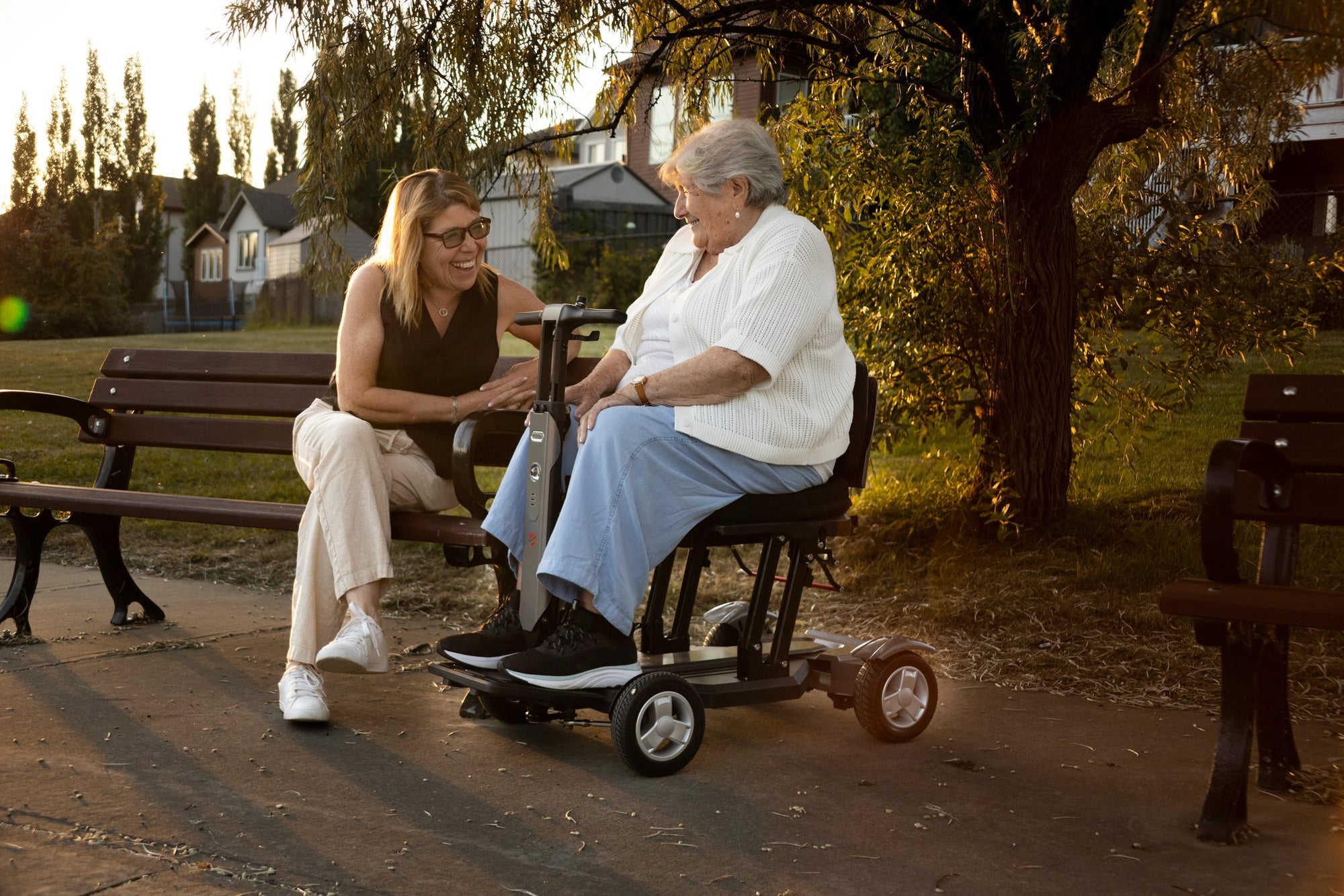 Older woman riding the Travel Buggy Helix mobility scooter and chatting with a companion on a park bench at sunset
